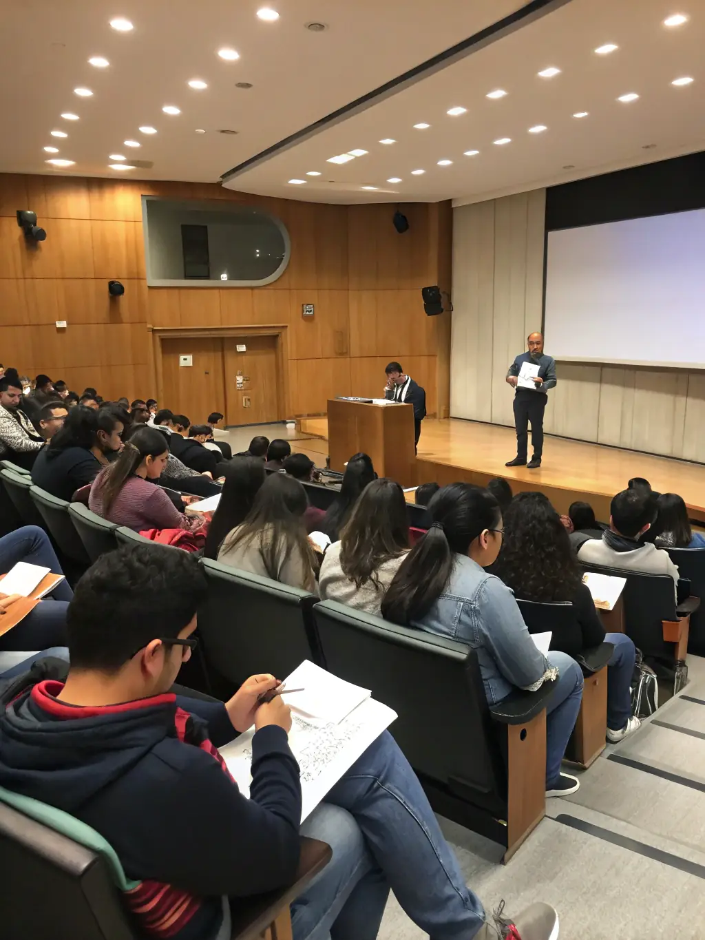 A group of diverse first-time homebuyers attentively listening to Garrett during a home buying seminar, with presentation slides visible in the background.